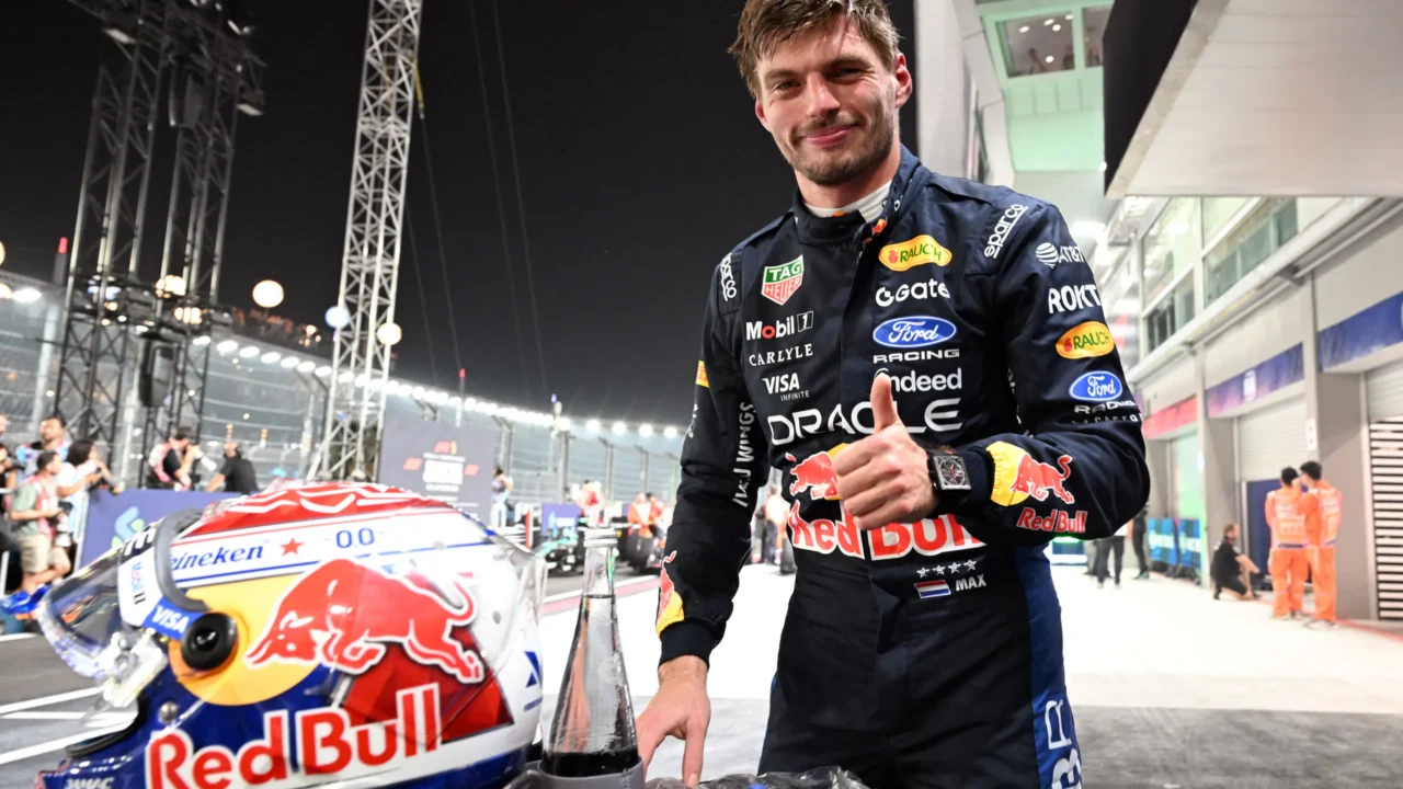 Piloto de macacão azul-marinho com patrocínios da Red Bull faz sinal de positivo ao lado de capacete estampado com o touro da equipe, em box iluminado à noite