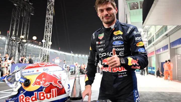 Piloto de macacão azul-marinho com patrocínios da Red Bull faz sinal de positivo ao lado de capacete estampado com o touro da equipe, em box iluminado à noite