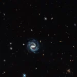 Uma galáxia espiral sinuosa (centro) aparece no mesmo campo de visão que uma vívida lente gravitacional (canto direito) nesta imagem do Hubble. A lente é composta por uma galáxia elíptica avermelhada em primeiro plano e pelos arcos azuis de uma galáxia ao fundo, ampliada.