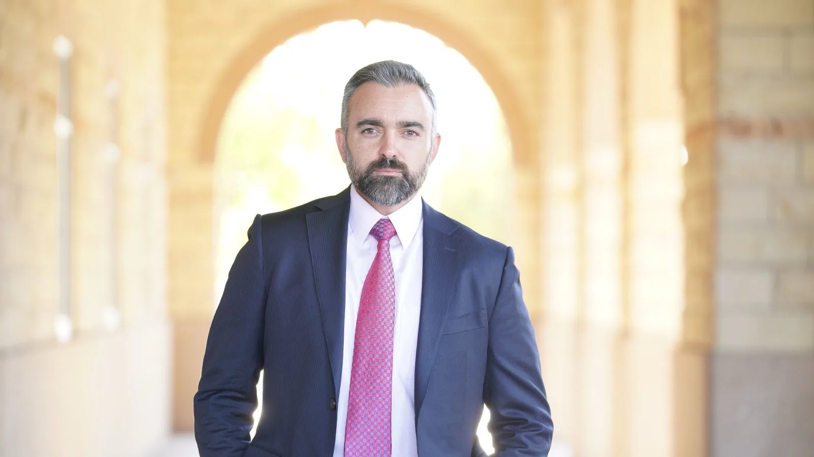 O procurador-geral do Novo México, Raúl Torrez, de terno azul e gravata rosa, em um corredor com arcos