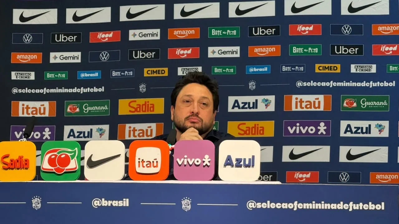Técnico de camisa escura, com a mão no queixo, fala em coletiva de imprensa diante de painéis de patrocinadores da Seleção Feminina