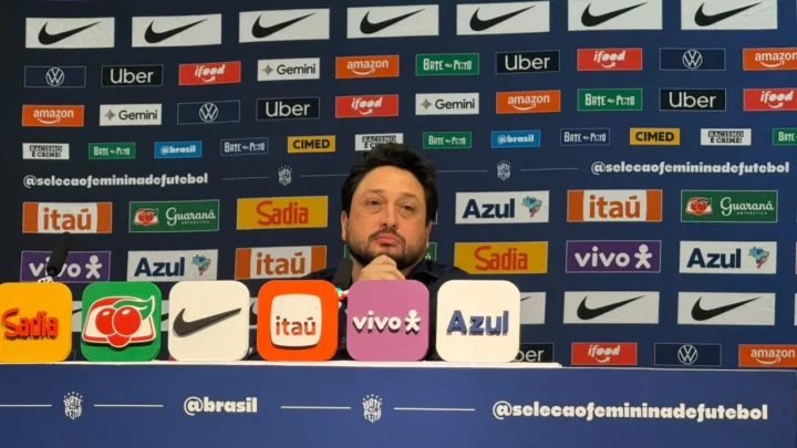 Técnico de camisa escura, com a mão no queixo, fala em coletiva de imprensa diante de painéis de patrocinadores da Seleção Feminina