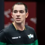 Atleta de camisa preta e verde, com patrocínio Tibhar e logo da CBTM, olha para o lado durante evento esportivo