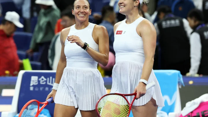 Duas tenistas de roupas brancas sorriem enquanto seguram raquetes, com torcida ao fundo em uma quadra de torneio