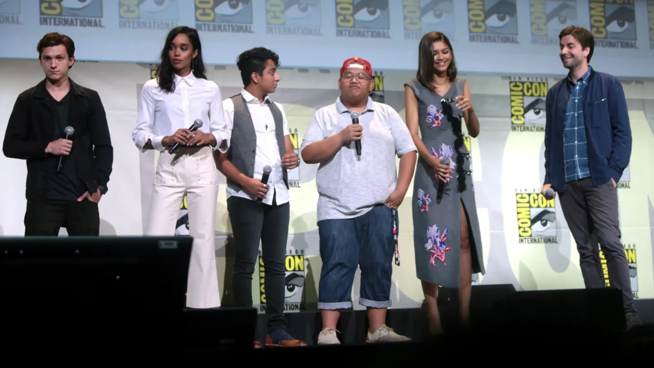 Seis participantes de painel sobre filme de super-heróis posam no palco da Comic-Con International com microfones em mãos