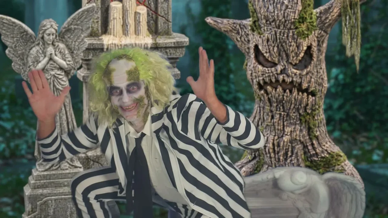 Personagem caracterizado como Beetlejuice, de terno listrado e cabelo verde, acena num cenário de cemitério, entre uma estátua de anjo e uma árvore assustadora