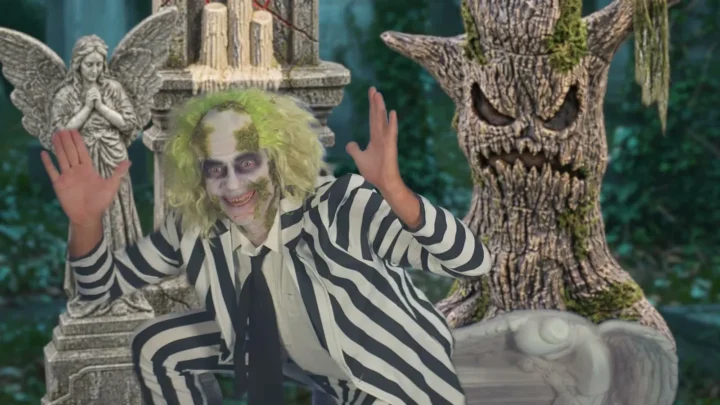 Personagem caracterizado como Beetlejuice, de terno listrado e cabelo verde, acena num cenário de cemitério, entre uma estátua de anjo e uma árvore assustadora