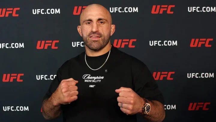 Homem careca e barbudo, de camiseta preta com a inscrição "Champion Mentality", faz gesto com os punhos cerrados diante de um painel com logos do UFC