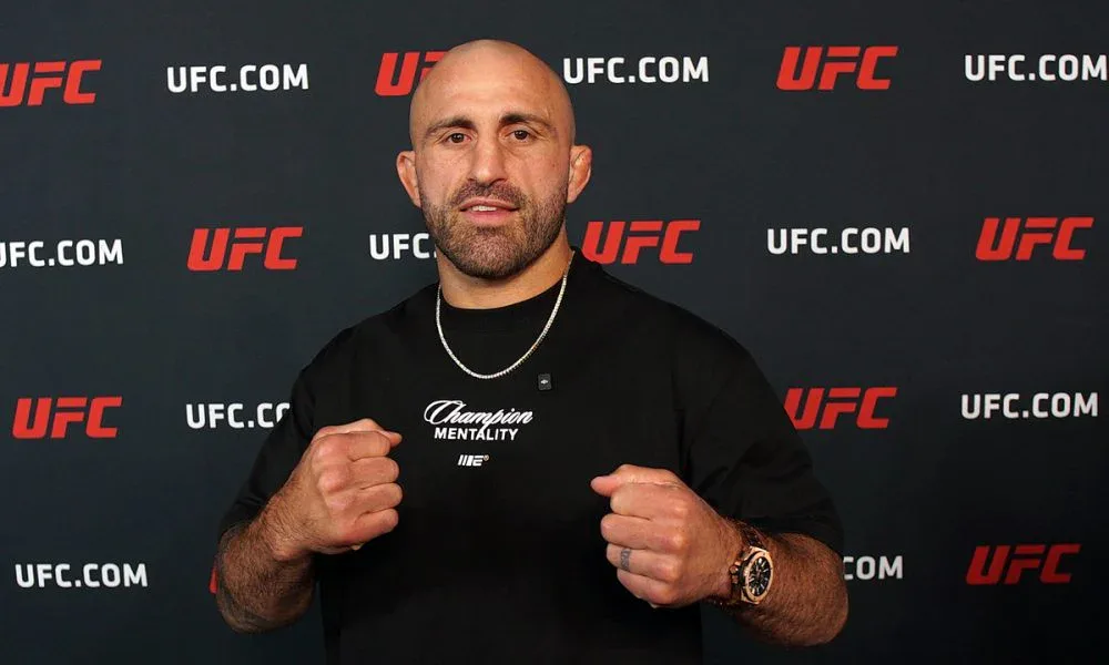 Homem careca e barbudo, de camiseta preta com a inscrição "Champion Mentality", faz gesto com os punhos cerrados diante de um painel com logos do UFC