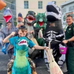 Criança fantasiada de dinossauro recebe doce de agente do Orange County Sheriff's Office durante evento comunitário de Halloween em Orlando. A edição 2026 do Trunk-or-Treat acontece na sexta-feira, 23 de outubro, das 18h às 20h, na sede de Operações Centrais (2500 W. Colonial Drive), com 12 estações