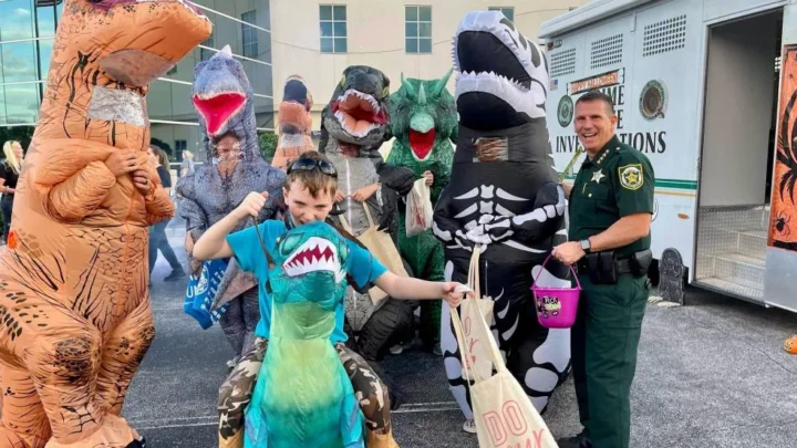 Criança fantasiada de dinossauro recebe doce de agente do Orange County Sheriff's Office durante evento comunitário de Halloween em Orlando. A edição 2026 do Trunk-or-Treat acontece na sexta-feira, 23 de outubro, das 18h às 20h, na sede de Operações Centrais (2500 W. Colonial Drive), com 12 estações