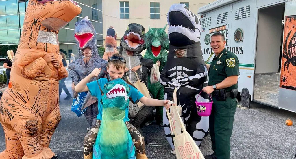 Criança fantasiada de dinossauro recebe doce de agente do Orange County Sheriff's Office durante evento comunitário de Halloween em Orlando. A edição 2026 do Trunk-or-Treat acontece na sexta-feira, 23 de outubro, das 18h às 20h, na sede de Operações Centrais (2500 W. Colonial Drive), com 12 estações