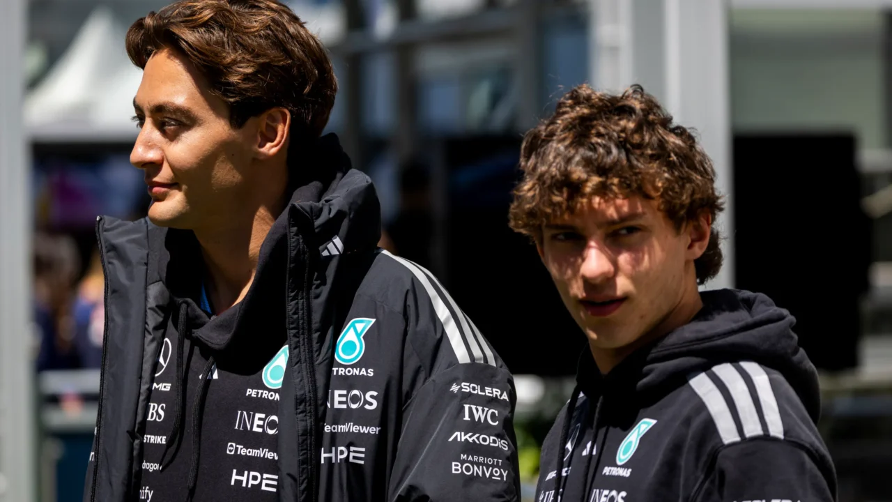 Dois pilotos da Mercedes em uniforme da equipe com patrocínios em destaque observam o cenário ao redor em ambiente de paddock