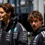 Dois pilotos da Mercedes em uniforme da equipe com patrocínios em destaque observam o cenário ao redor em ambiente de paddock