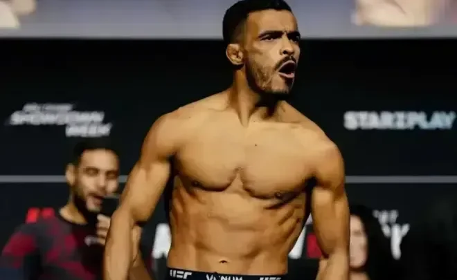 Lutador sem camisa, de bermuda preta com a marca UFC, posa de boca aberta em frente a um painel com patrocinadores durante pesagem de evento de MMA