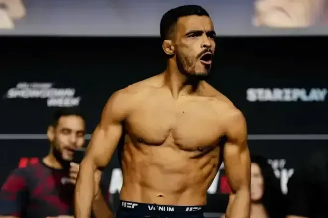 Lutador sem camisa, de bermuda preta com a marca UFC, posa de boca aberta em frente a um painel com patrocinadores durante pesagem de evento de MMA