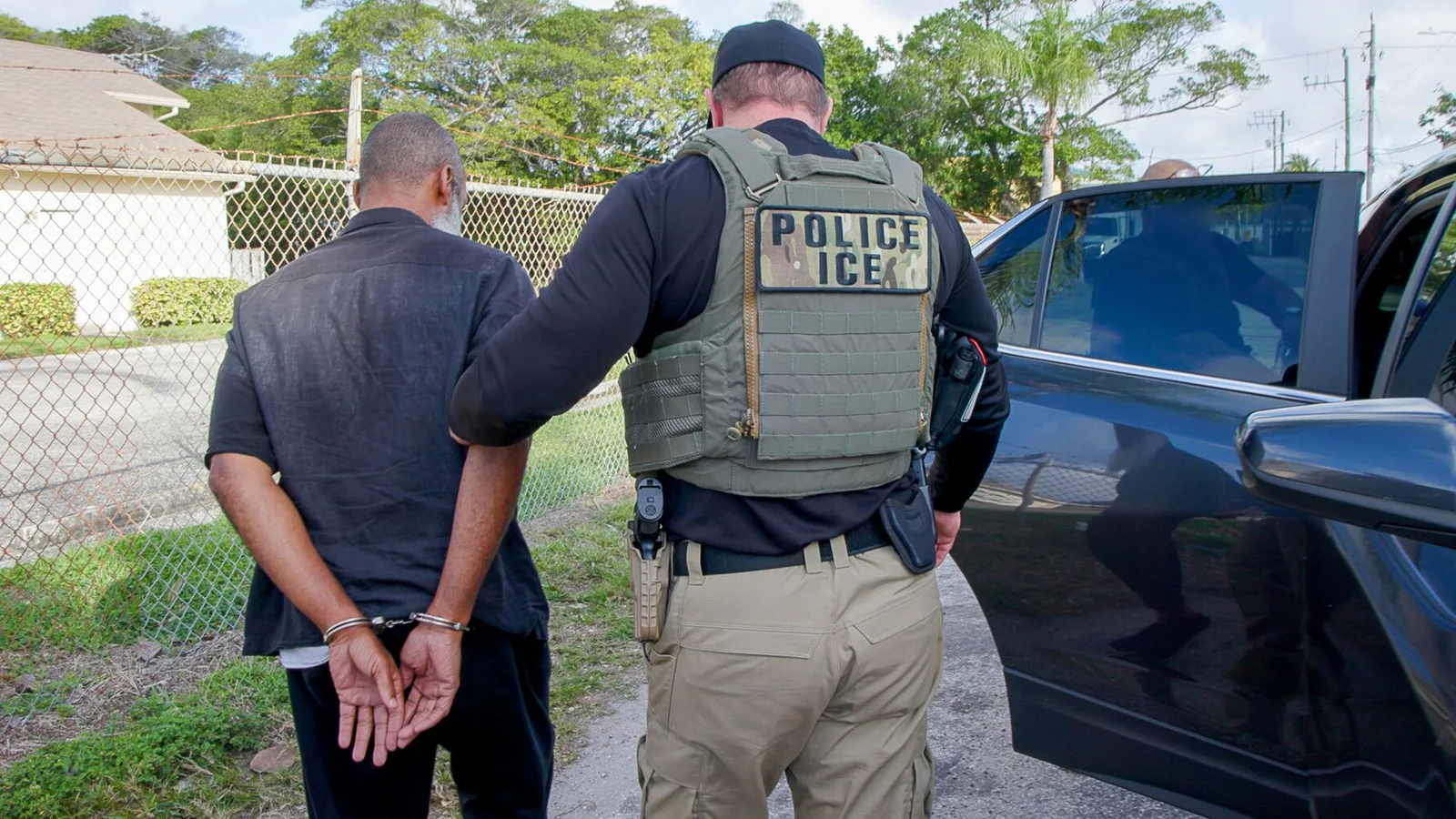 Agente do ICE, de costas, com colete escrito "Police ICE", conduz um homem algemado até um carro durante uma operação em West Palm Beach, na Flórida, em fevereiro de 2025; foto de arquivo, sem relação com o caso