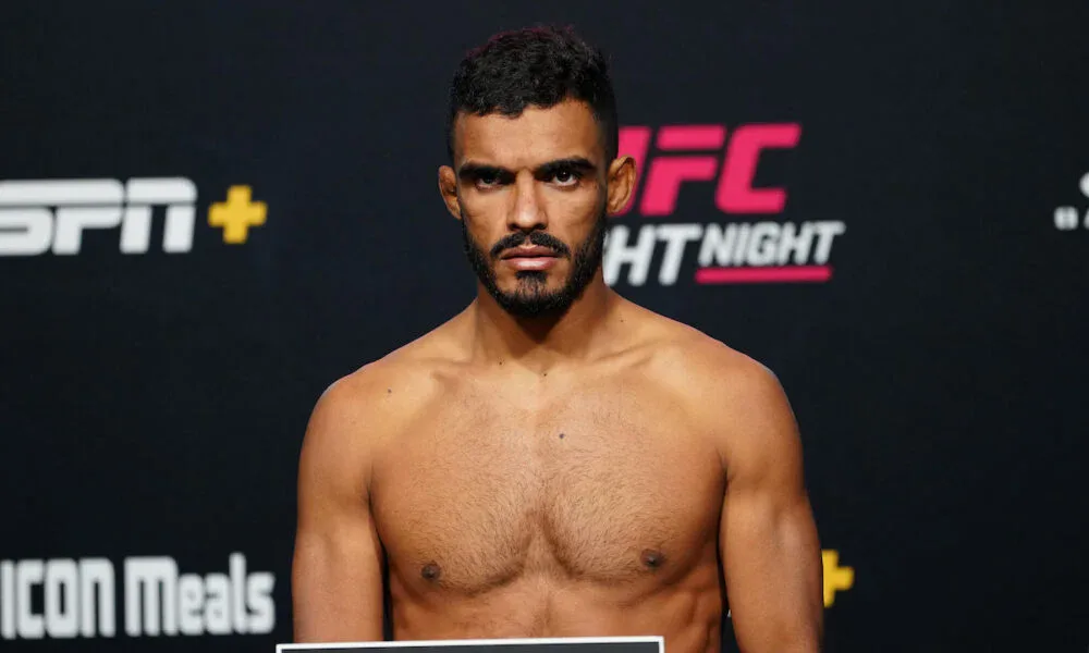 Homem de peito nu, com barba e cabelo escuro, posa em frente a um painel de patrocinadores do UFC Fight Night e da ESPN+ durante a pesagem
