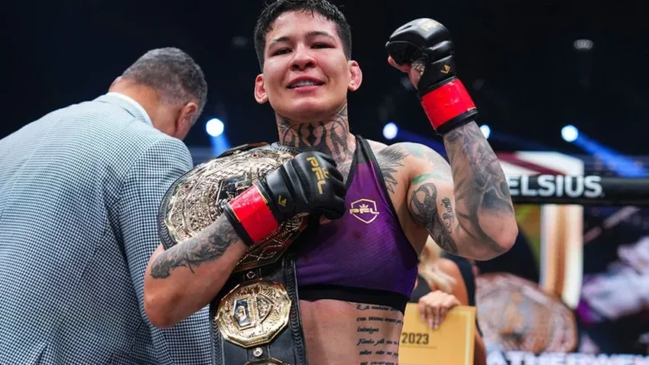 Lutadora de MMA com roupa roxa e luvas pretas e vermelhas, com cinturão dourado no ombro, comemora no ringue com o braço erguido e sorriso no rosto