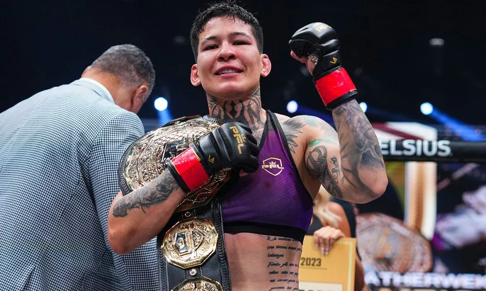 Lutadora de MMA com roupa roxa e luvas pretas e vermelhas, com cinturão dourado no ombro, comemora no ringue com o braço erguido e sorriso no rosto
