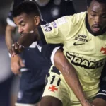 Jogador de camisa amarela do Vasco, com escudo e patrocínios visíveis, disputa a bola com adversário de uniforme preto em lance dentro de campo à noite
