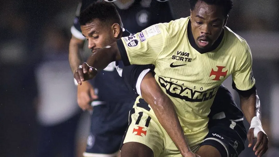 Jogador de camisa amarela do Vasco, com escudo e patrocínios visíveis, disputa a bola com adversário de uniforme preto em lance dentro de campo à noite