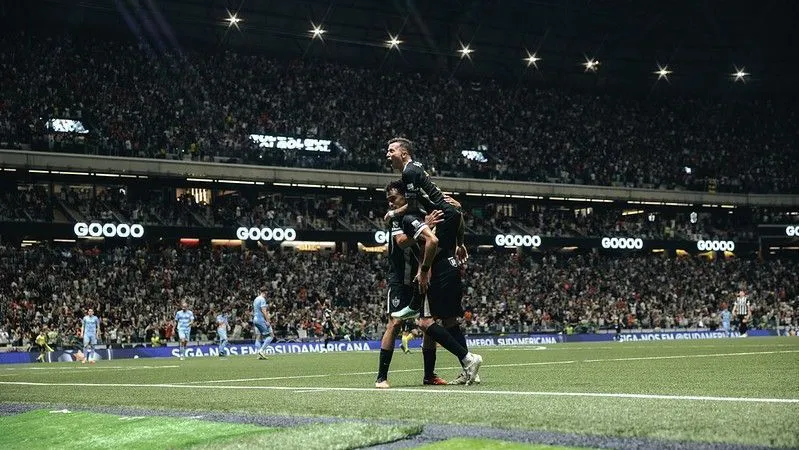 Jogadores do Atlético-MG comemoram um gol em estádio lotado, com o público nas arquibancadas e placas de patrocínio ao redor do gramado