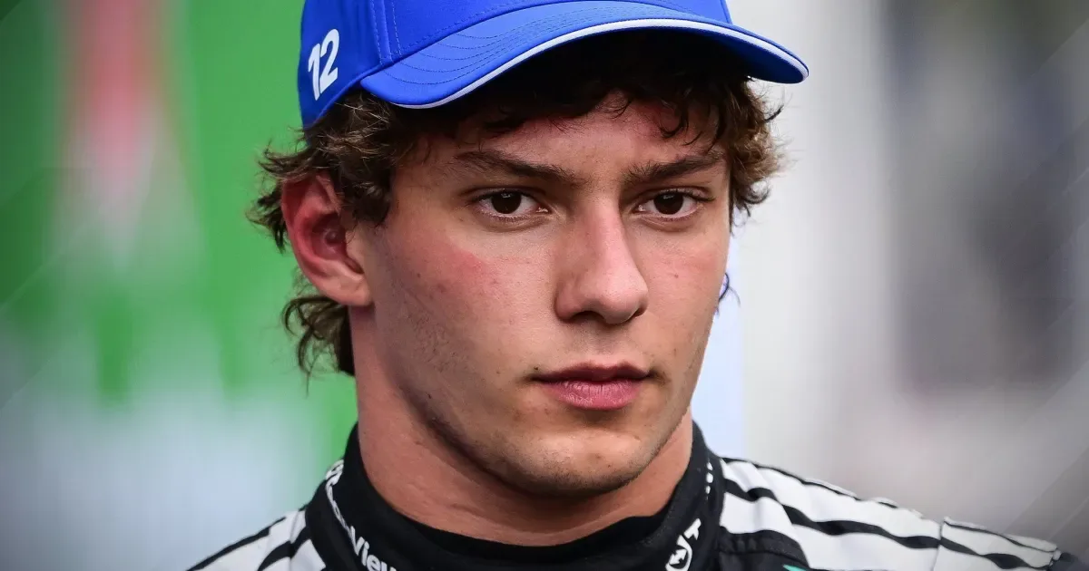 Jovem piloto de macacão preto e branco da equipe, usando boné azul com o número 12, com expressão séria e olhar voltado para a lateral