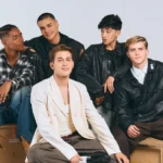 Cinco integrantes do grupo Santos Bravos posam juntos sentados e em pé diante de fundo neutro, usando roupas em tons de preto, branco e tons naturais