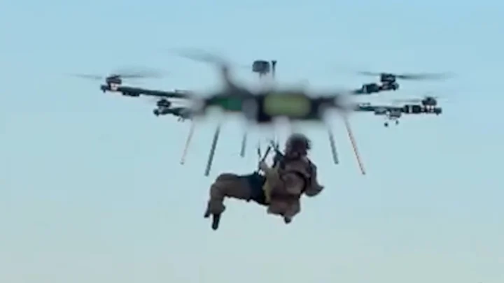 Soldado ferido voa suspenso sob um drone de seis hélices, sobre um campo de vegetação seca, durante a retirada da linha de frente