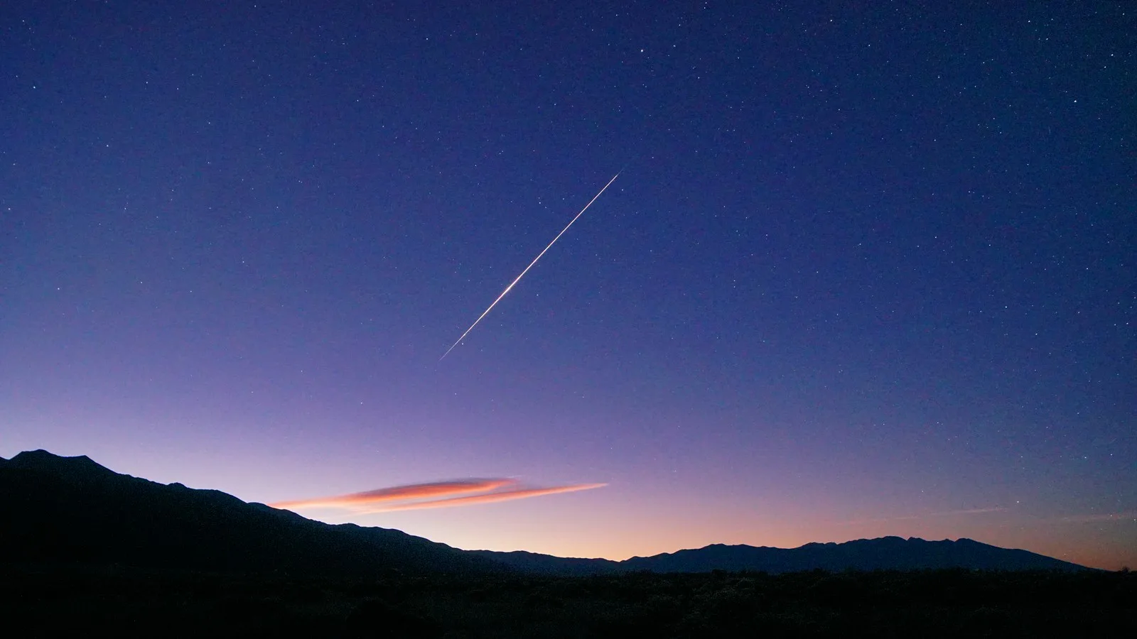 Meteoro das Orionídeas risca o céu do amanhecer sobre montanhas em silhueta, com uma nuvem lenticular alongada e alaranjada perto do horizonte, em outubro de 2025