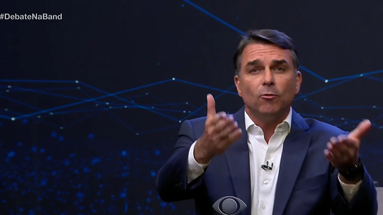 Flávio Bolsonaro, de terno azul e camisa branca, gesticula com as duas mãos durante o 3º bloco