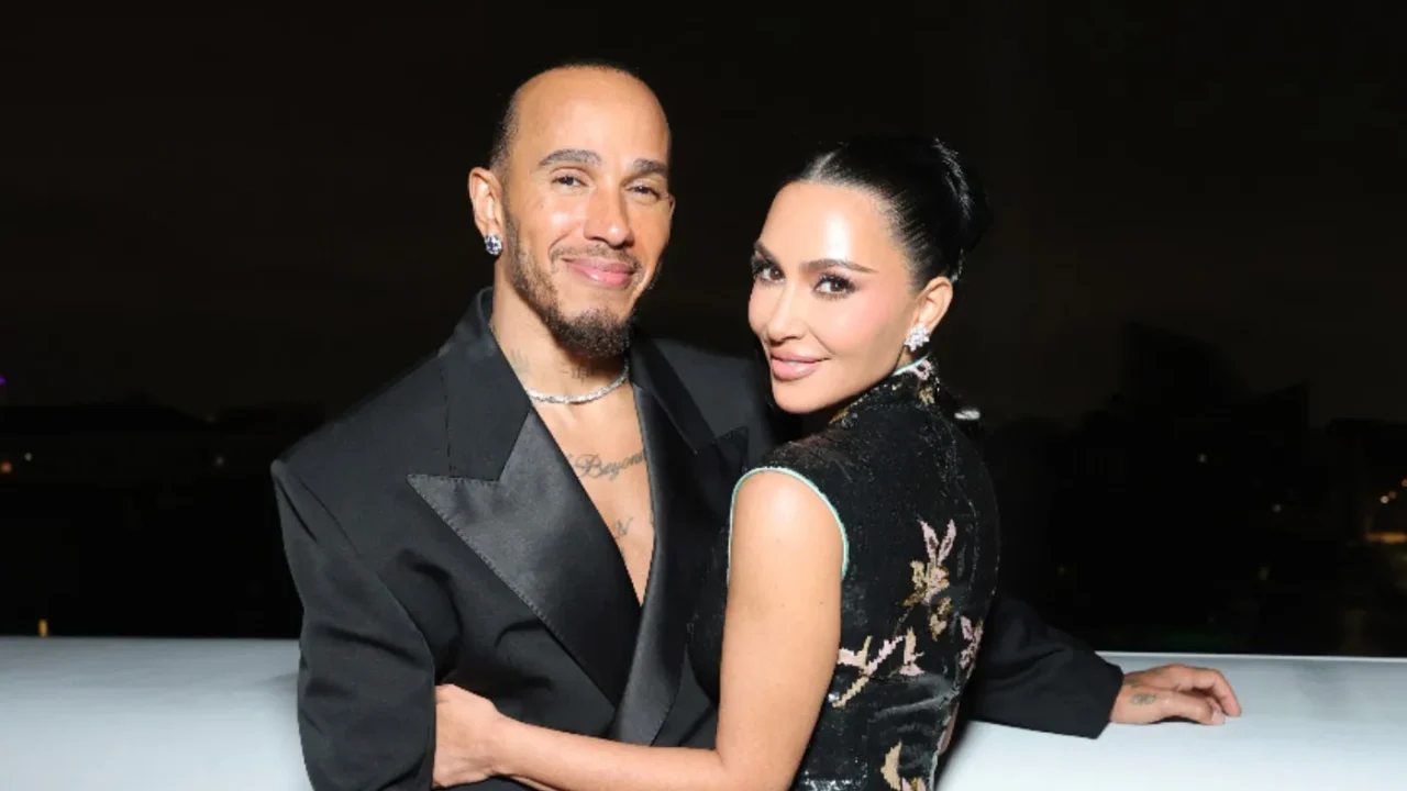 Dois pessoas posam juntas em evento noturno, com um homem usando blazer preto sobre fundo escuro e uma mulher em vestido preto com detalhes dourados