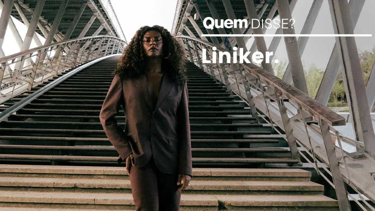 Mulher com cabelo comprido cacheado veste blazer marrom escuro em pé em escadaria de estrutura metálica