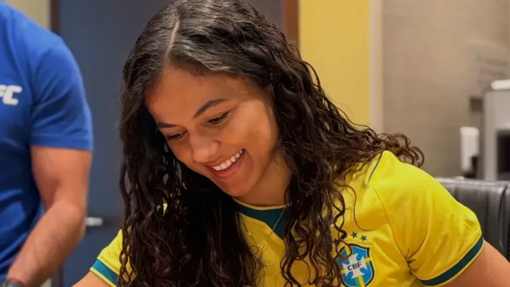 Mulher usando camiseta amarela da seleção brasileira sorri durante encontro em ambiente interno com outra pessoa ao fundo vestindo azul