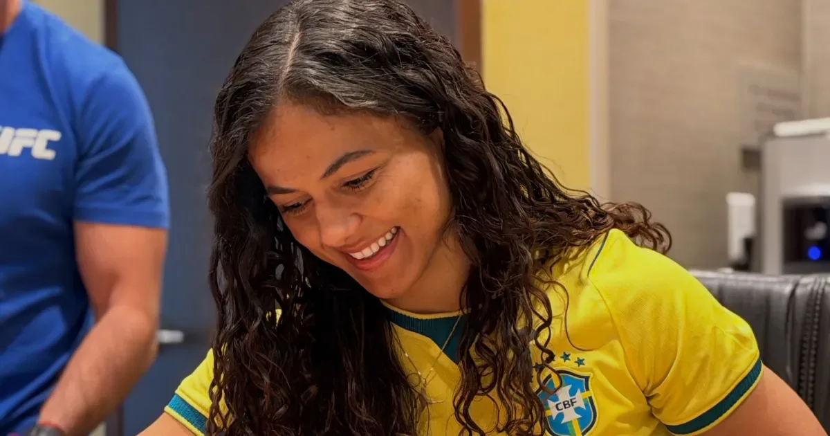 Mulher usando camiseta amarela da seleção brasileira sorri durante encontro em ambiente interno com outra pessoa ao fundo vestindo azul