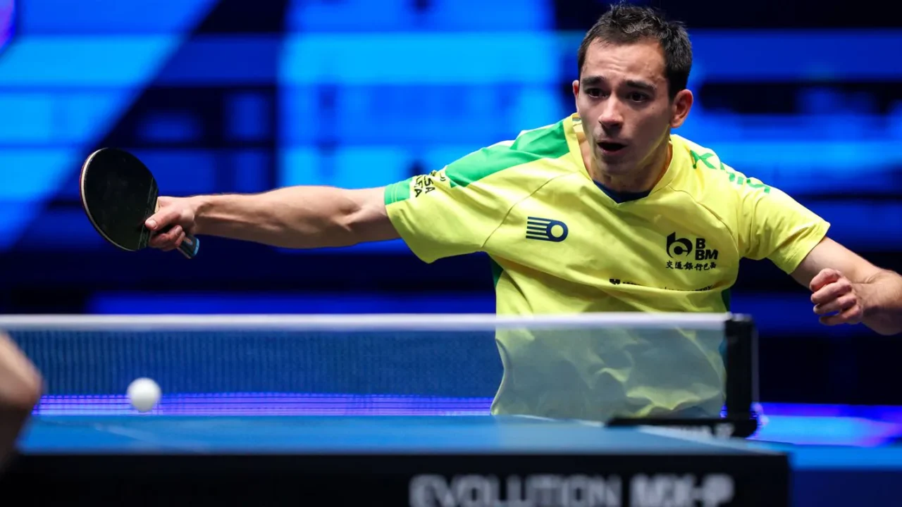 Atleta de tênis de mesa em uniforme amarelo-neon executa saque durante partida em competição internacional com iluminação azul ao fundo