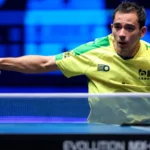 Atleta de tênis de mesa em uniforme amarelo-neon executa saque durante partida em competição internacional com iluminação azul ao fundo