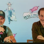 Dois homens seguram microfones durante painel em evento, com cenário ao fundo estampado com personagens de ficção científica e naves espaciais