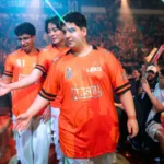 Jogadores com camisas laranja da LOS cumprimentam a torcida ao entrar em uma arena lotada, com luzes coloridas e plateia com bastões de LED