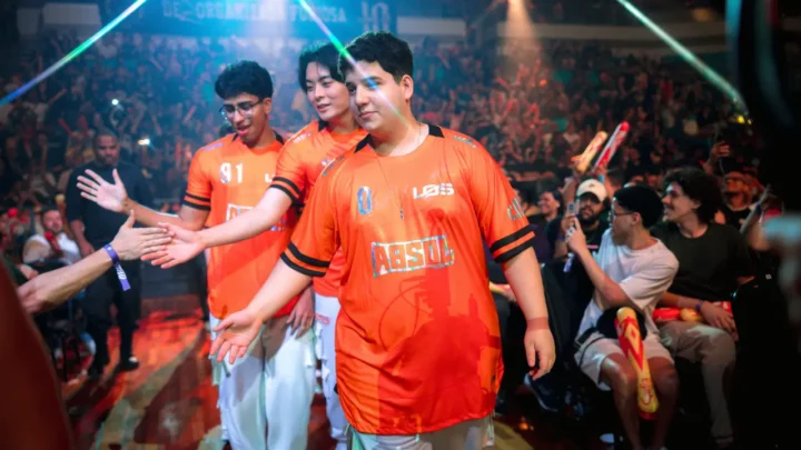 Jogadores com camisas laranja da LOS cumprimentam a torcida ao entrar em uma arena lotada, com luzes coloridas e plateia com bastões de LED