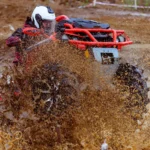 Piloto de capacete branco passa com um quadriciclo vermelho por uma poça e levanta uma parede de lama numa prova de off-road na Rússia, em 2020 (imagem ilustrativa)
