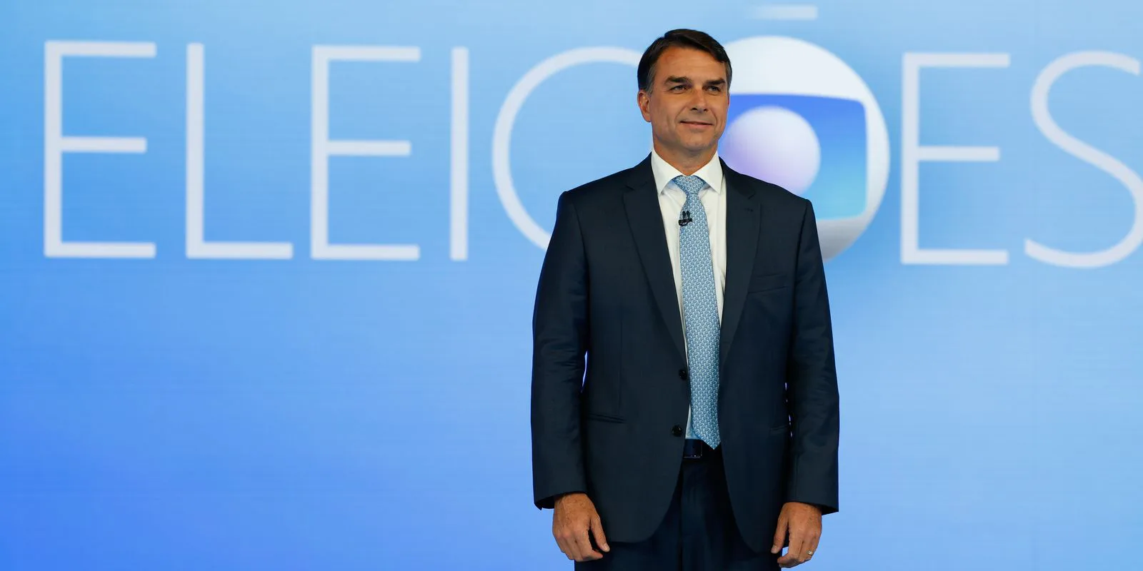 Flávio Bolsonaro, sorrindo, de terno azul-marinho e gravata azul-clara, no cenário do debate da TV Globo, no Rio de Janeiro, em 23 de outubro de 2026