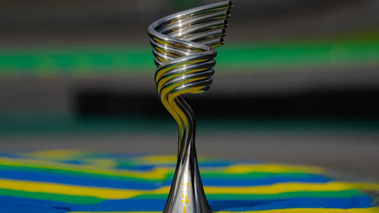 Troféu prateado em espiral, com detalhes dourados na base, posado sobre piso pintado nas cores verde, amarelo e azul