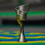 Troféu prateado em espiral, com detalhes dourados na base, posado sobre piso pintado nas cores verde, amarelo e azul