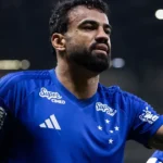 Jogador de camisa azul do Cruzeiro, com barba, faz gesto com a mão levantada e três dedos abertos durante partida à noite em estádio
