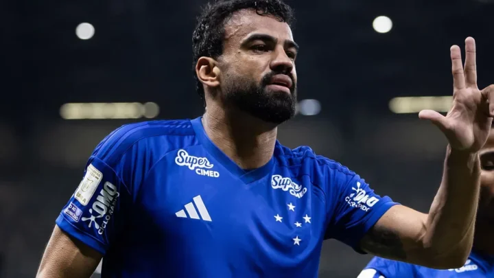 Jogador de camisa azul do Cruzeiro, com barba, faz gesto com a mão levantada e três dedos abertos durante partida à noite em estádio