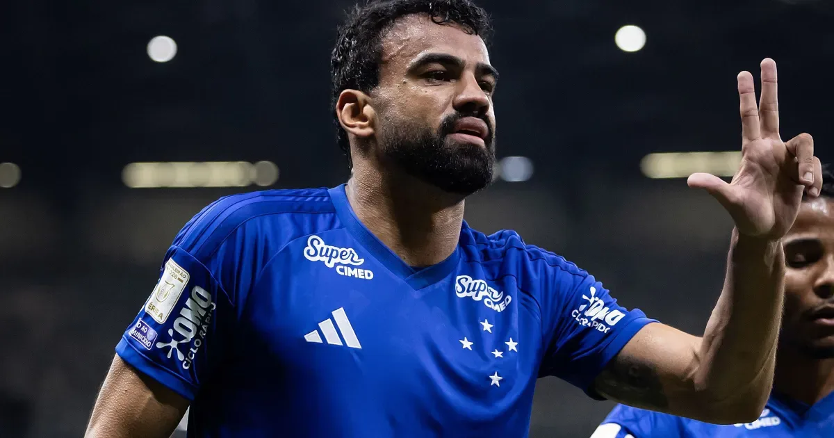 Jogador de camisa azul do Cruzeiro, com barba, faz gesto com a mão levantada e três dedos abertos durante partida à noite em estádio