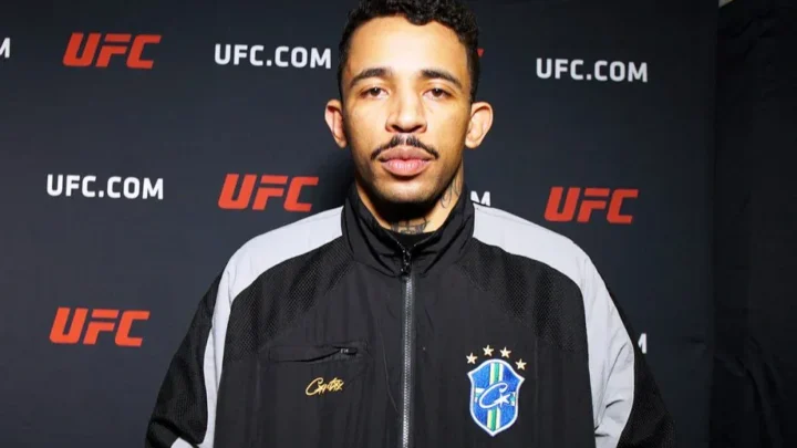 Homem de jaqueta preta com detalhes cinza e um escudo azul com a bandeira do Brasil, posando diante de um painel com o logotipo do UFC