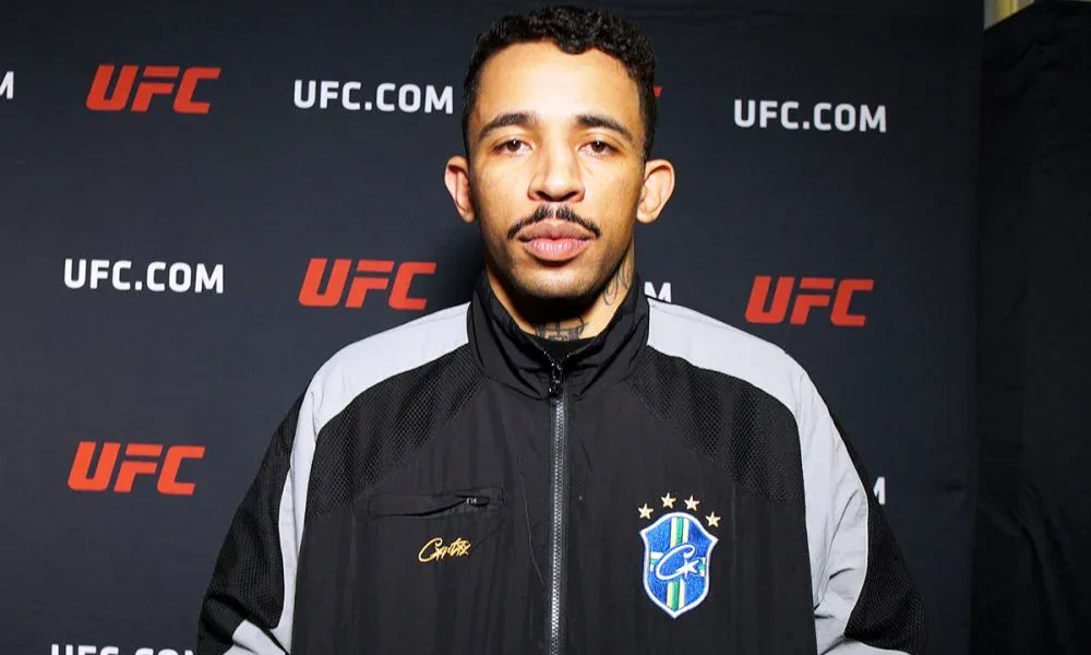 Homem de jaqueta preta com detalhes cinza e um escudo azul com a bandeira do Brasil, posando diante de um painel com o logotipo do UFC